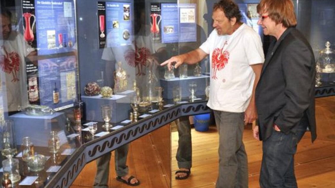 Zwei Männer stehen vor Vitrinen in einem Museum und betrachten verschiedene Objekte. Einer der Männer zeigt auf ein Objekt, während der andere interessiert zuhört. | © Jakobihaus Soboth