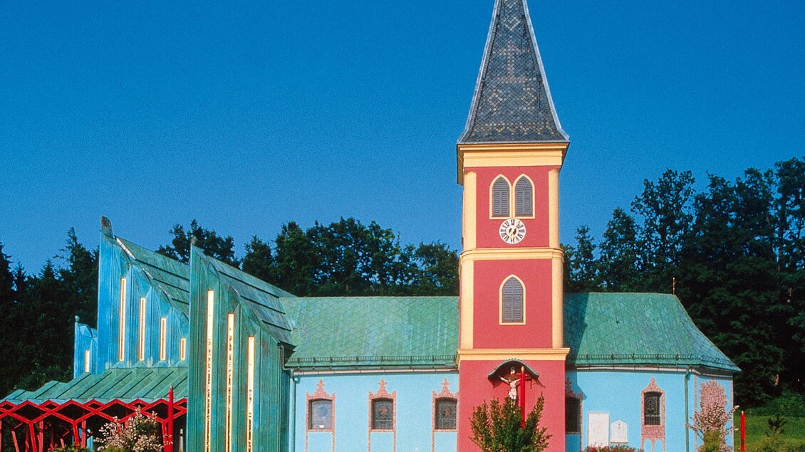 Eine bunte Kirche mit einem blauen Dach und einem hohen, spitzen Turm. Die Fassade ist in lebhaften Farben gestaltet und von Bäumen umgeben. | © Hans Wiesenhofer