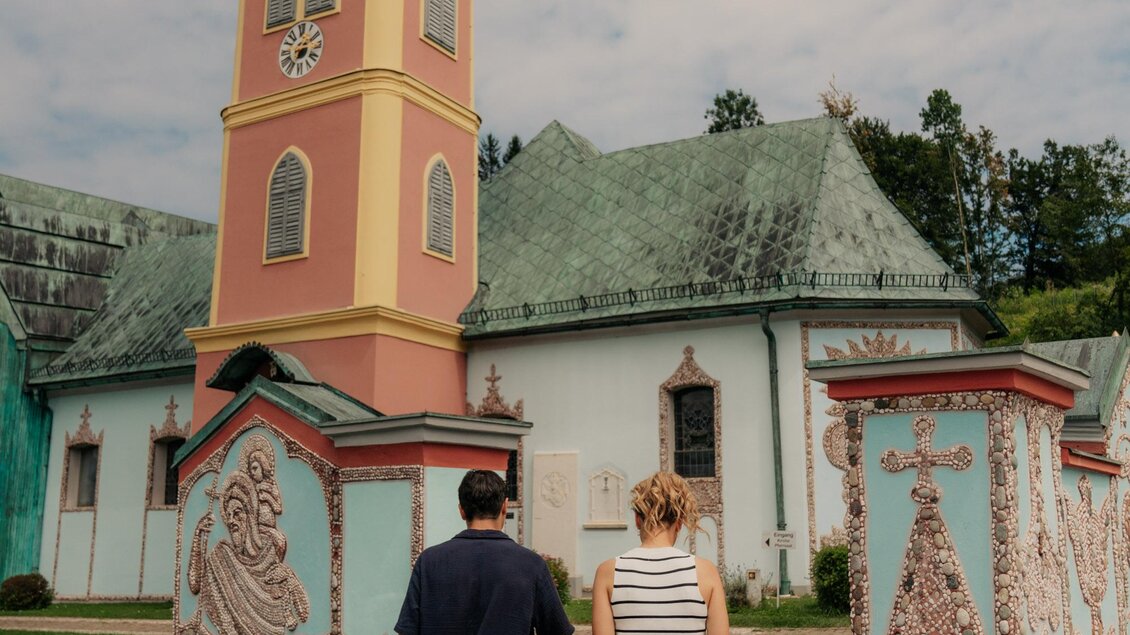 Ein Paar geht auf einem Weg zu einem historischen Gebäude mit einer bunten Fassade und einem hohen Turm. Der Himmel ist bewölkt und die Umgebung ist grün. | © (c) Region Graz - studio draussen