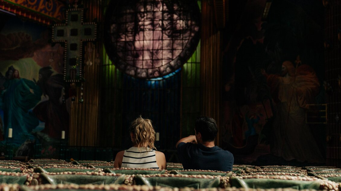 Zwei Personen sitzen auf Bänken in einem alten Kino. Im Hintergrund ist ein großes, buntes Fenster mit Bildern zu sehen. | © (c) Region Graz - studio draussen