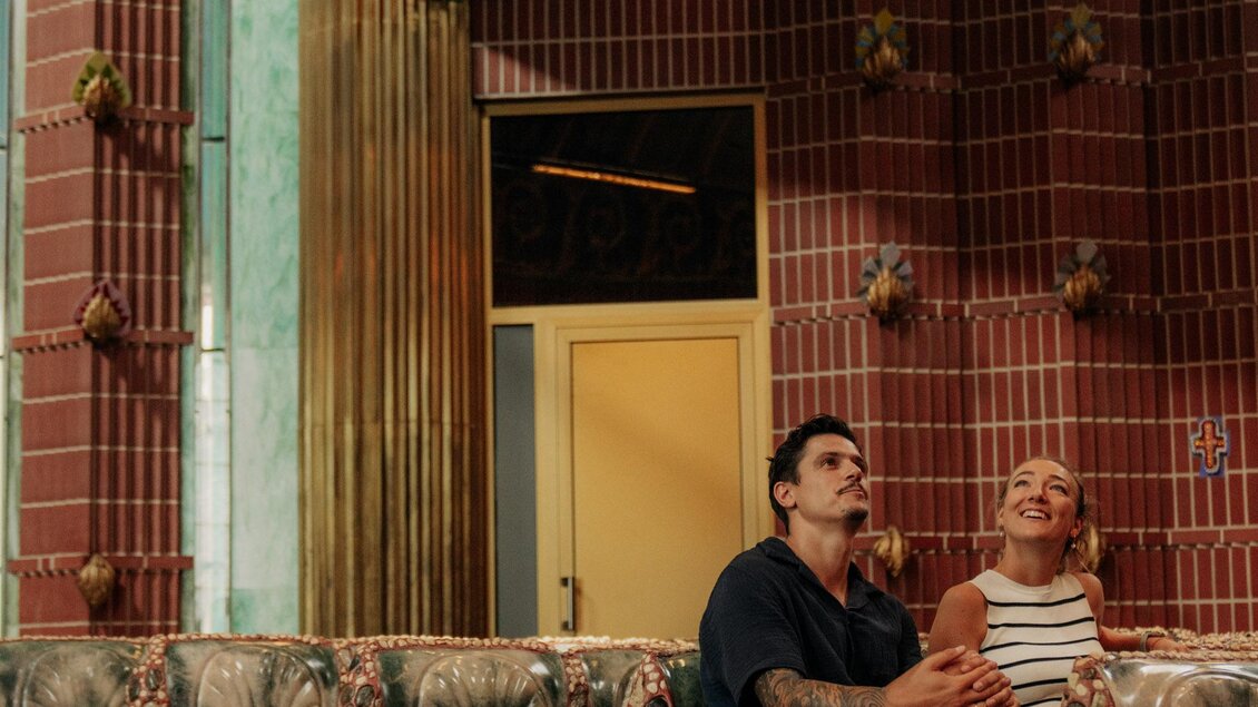 Ein gemütlicher Innenraum mit bunten Möbeln und kreativer Dekoration. Zwei Personen sitzen entspannt auf einem Sofa und unterhalten sich. | © (c) Region Graz - studio draussen