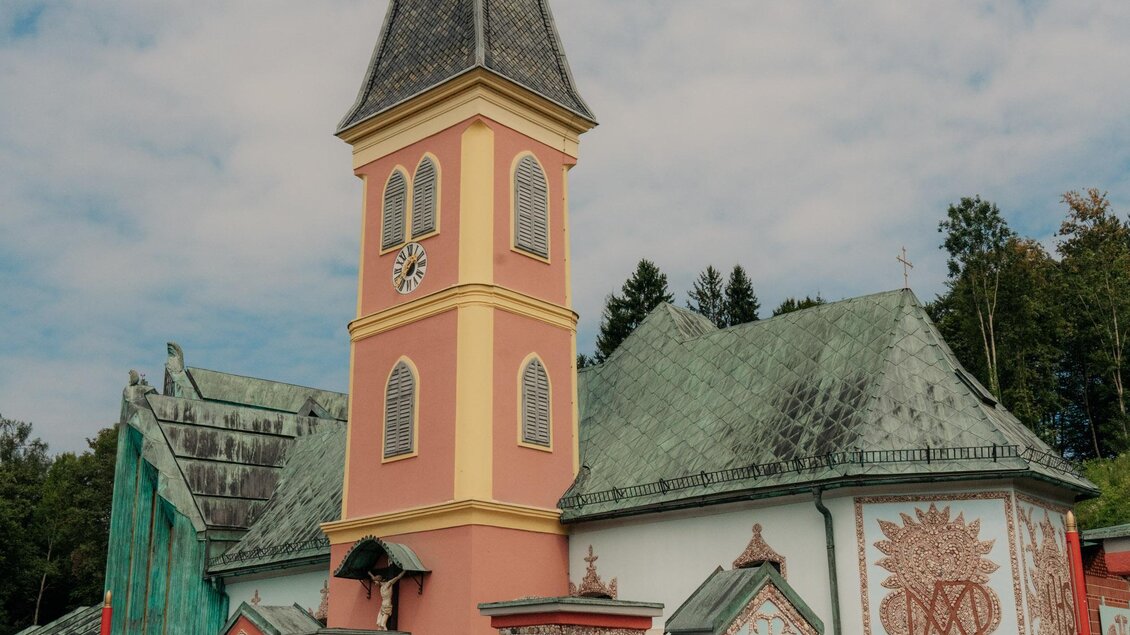 Eine farbenfrohe Kirche mit einem hohen Turm und schönen Wandmalereien. Die Umgebung ist grün und einladend. | © (c) Region Graz - studio draussen