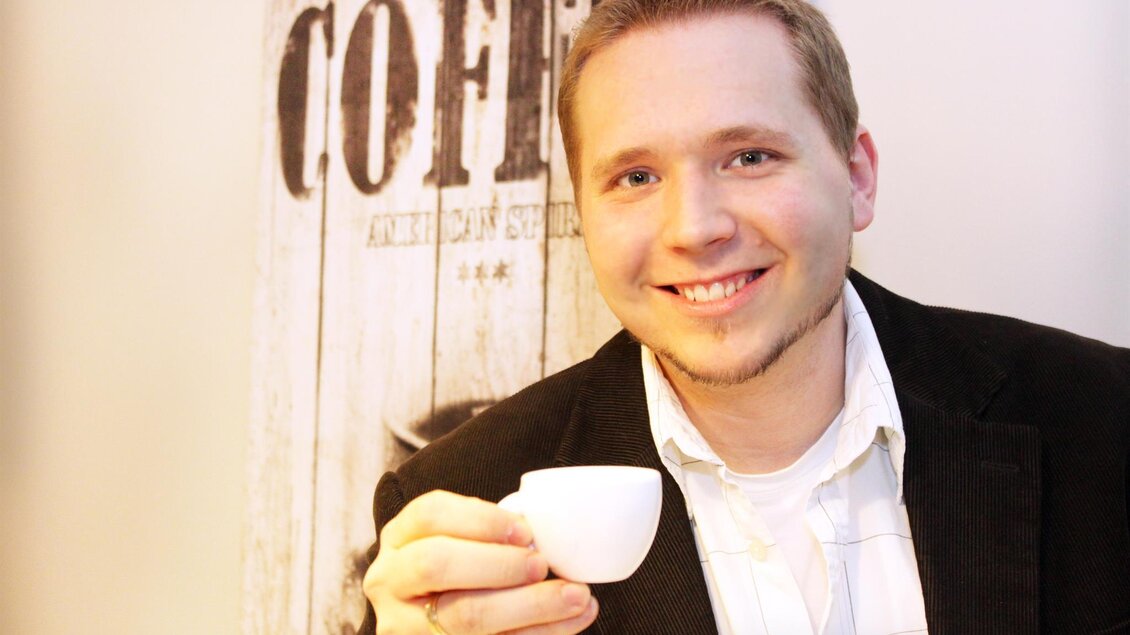 Ein lachender Mann hält eine kleine Tasse in der Hand. Im Hintergrund ist ein Kaffeethema sichtbar. | © Jamila Coffee GmbH