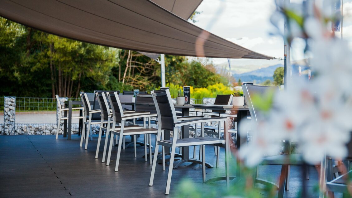 Ein modern eingerichteter Außenbereich mit Tischen und Stühlen unter einem Sonnenschutz. Im Hintergrund sind grüne Bäume und eine schöne Landschaft zu sehen. | © Jamila Coffee GmbH