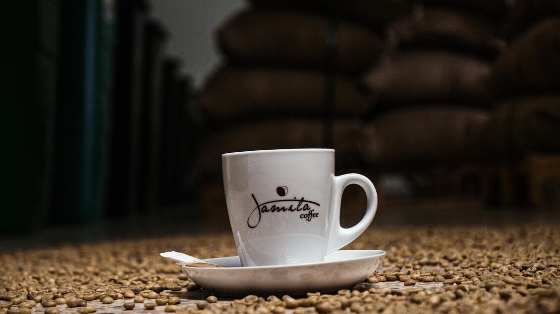 Eine Kaffeetasse steht auf dem Boden, umgeben von Kaffeebohnen. Im Hintergrund sind Säcke mit geröstetem Kaffee gestapelt. | © Jamila Coffee GmbH
