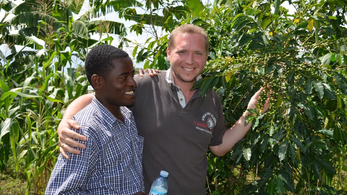 Zwei Männer stehen lächelnd vor einer Kaffeepflanze in einer grünen Umgebung. Der eine hält eine Wasserflasche in der Hand. | © Jamila Coffee GmbH