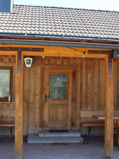 Ein traditionelles Holzhaus mit einem massiven Dach. Vor dem Haus befinden sich Holzbänke und Tische. | © Kogler`s Jausenhütt`n