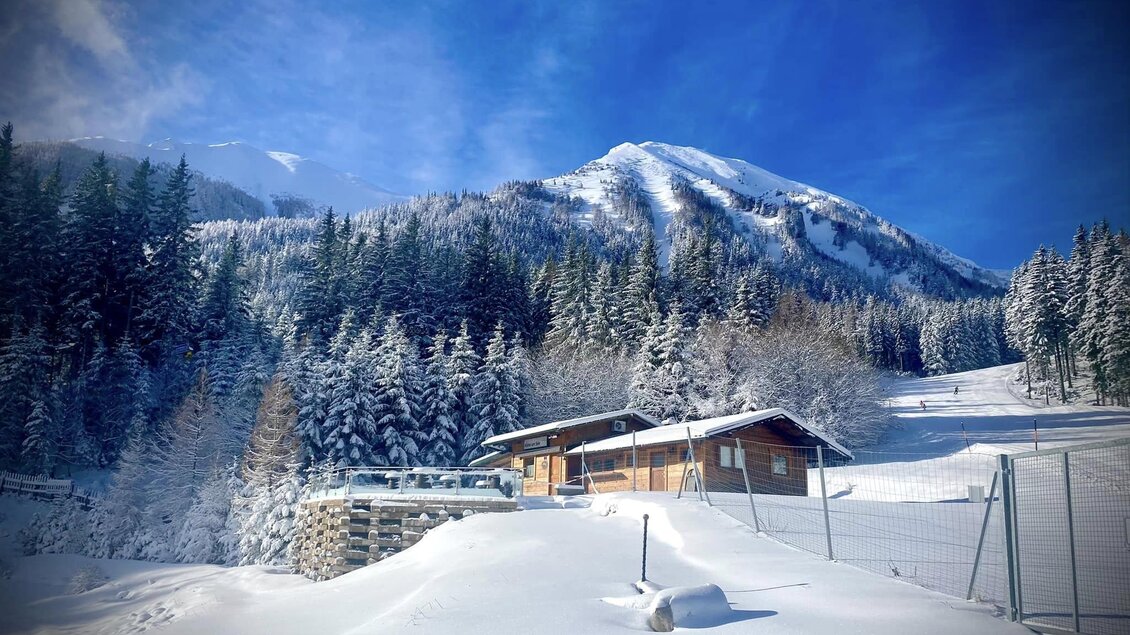 Eine malerische Winterlandschaft mit schneebedeckten Bergen und Tannenbäumen. Ein gemütliches Holzhaus liegt eingebettet im frischen Schnee. | © Marschnig