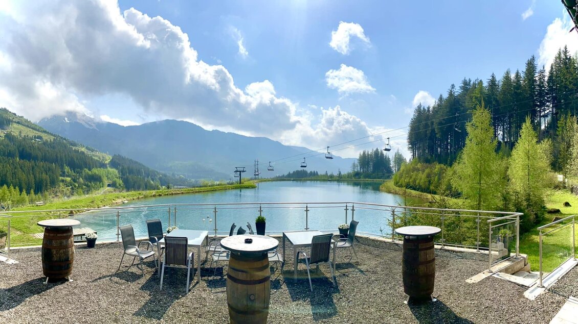 Ein malerischer Blick auf einen See umgeben von Bergen und Wäldern. Tische und Stühle laden zum Entspannen im Freien ein. | © Marschnig