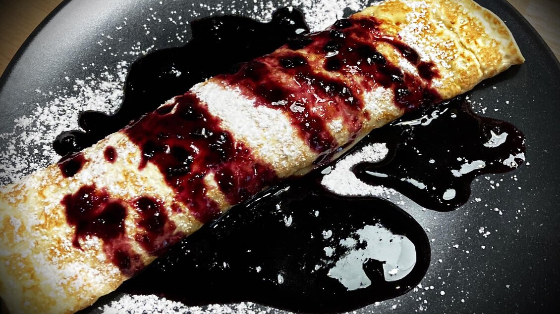 Ein leckerer Pfannkuchen mit Fruchtsoße und Puderzucker. Der Pfannkuchen liegt auf einem schwarzen Teller. | © Marschnig