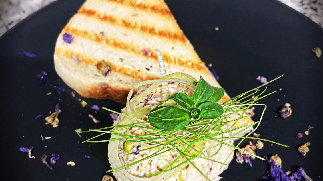 Ein Gericht mit einer cremigen Vorspeise, dekoriert mit Kräutern und essbaren Blüten. Dazu ein Stück geröstetes Brot auf einem schwarzen Teller. | © Marschnig