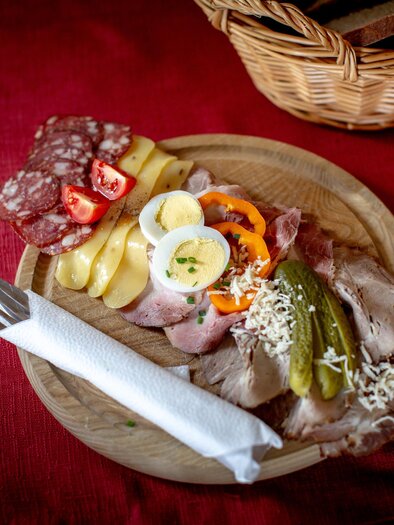 A wooden board with a selection of sausage, ham, cheese, and vegetables. Next to it is cutlery and a basket of bread in the background. | © Tourismusverband Murau