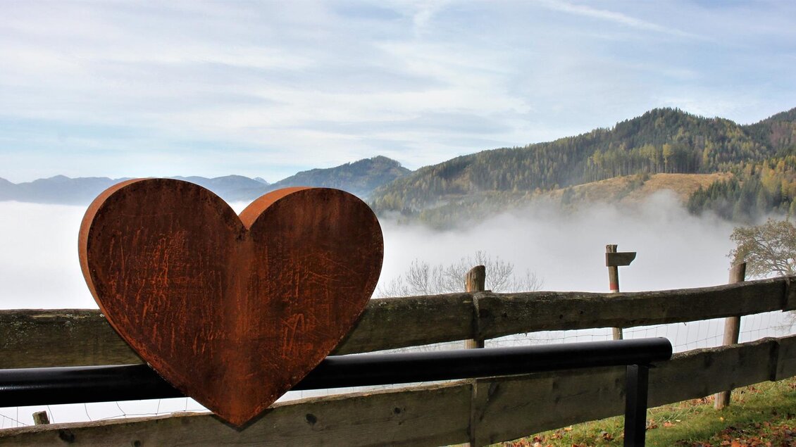 Ein großes Herz aus Holz steht vor einer malerischen Berglandschaft. Dahinter sind Nebel und sanfte Hügel zu sehen.