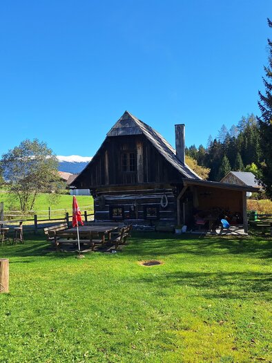 Ein traditionelles Haus in ländlicher Umgebung mit grünen Wiesen und Bäumen. Im Vordergrund befinden sich Holzmöbel und eine ruhige Atmosphäre. | © Jausenstation Russkeusche