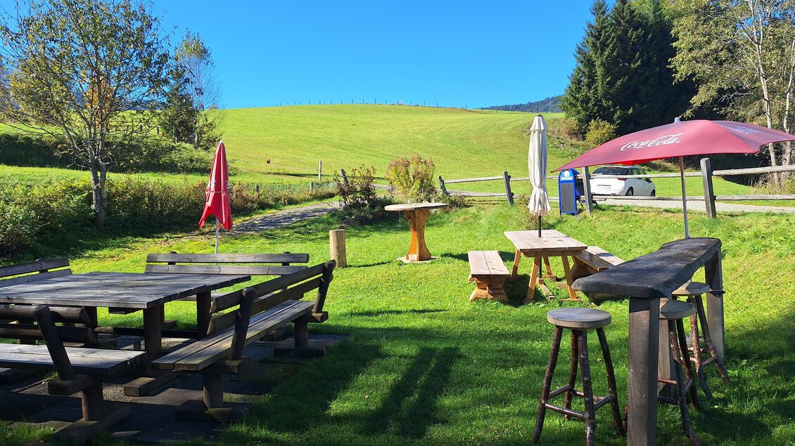 Eine einladende Grünfläche mit Holzmöbeln und Sonnenschirmen. Im Hintergrund erstreckt sich eine sanfte Wiese unter einem klaren blauen Himmel. | © Jausenstation Russkeusche