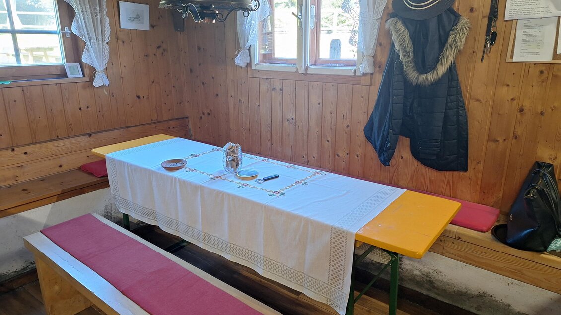 Ein rustikales Zimmer mit einer langen Tischbank und einem gedeckten Tisch. An der Wand hängen ein Hut und eine Jacke. | © Jausenstation Russkeusche