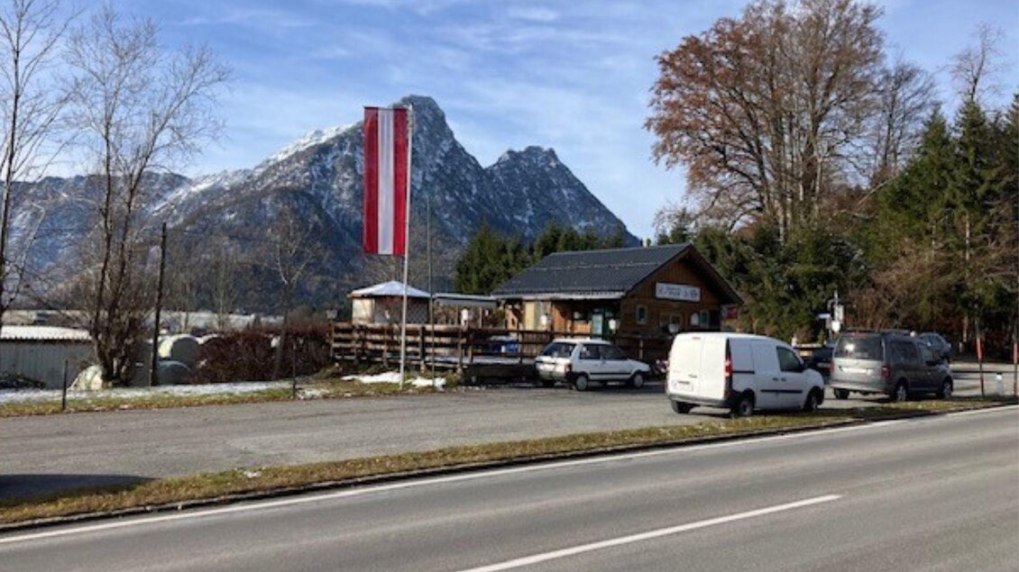 Ein Gebäude mit einer österreichischen Fahne in der Nähe einer Straße, umgeben von Bergen und Bäumen. Die Szenerie zeigt eine ruhige, ländliche Atmosphäre. | © TVB_Ausseerland Salzkammergut_Heike Temmel