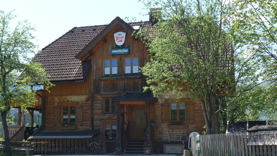 Ein traditionelles, holzverkleidetes Gasthaus umgeben von Bäumen. Die sonnige Umgebung und der einladende Eingang strahlen Gemütlichkeit aus. | © Familie Pliem