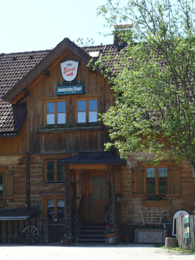 Ein traditionelles, holzverkleidetes Gasthaus umgeben von Bäumen. Die sonnige Umgebung und der einladende Eingang strahlen Gemütlichkeit aus. | © Familie Pliem