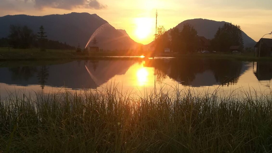 Ein wunderschöner Sonnenuntergang über einem ruhigen See, umgeben von Bergen. Die Sonne spiegelt sich im Wasser und schafft eine malerische Atmosphäre.