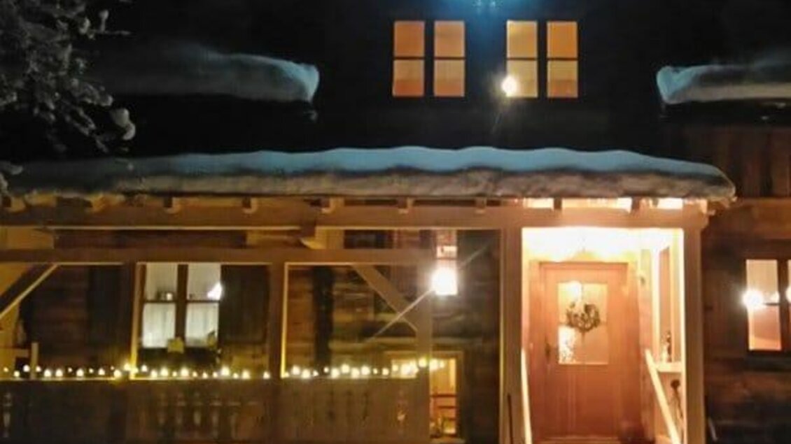 Ein gemütliches Holzhaus bei Nacht, beleuchtet von warmem Licht. Schneebedeckter Boden und festliche Dekorationen schaffen eine einladende Atmosphäre.