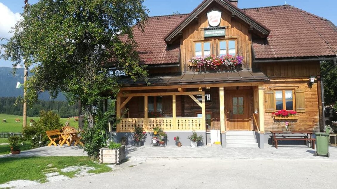 Ein charmantes Holzhaus mit einem Empfangsbereich und bunten Blumenkästen. Im Hintergrund sind grüne Wiesen und ein klarer Himmel zu sehen.
