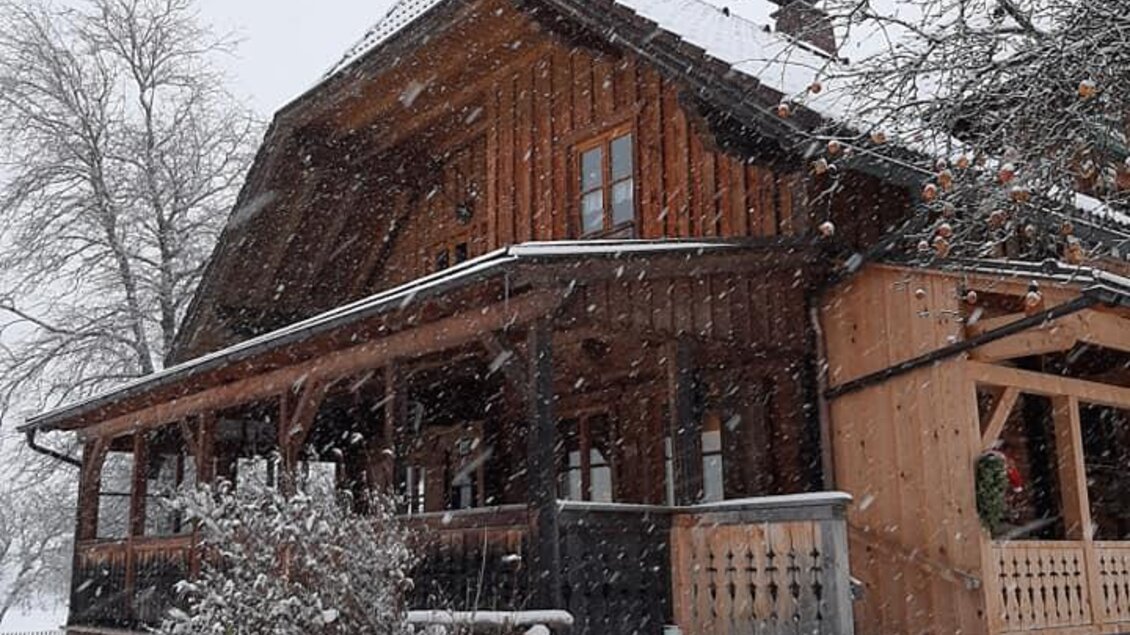 Ein charmantes Holzhaus im Schnee, umgeben von einem winterlichen Landschaftsbild. Großer Schnee fällt auf das Dach und den Vordereingang.