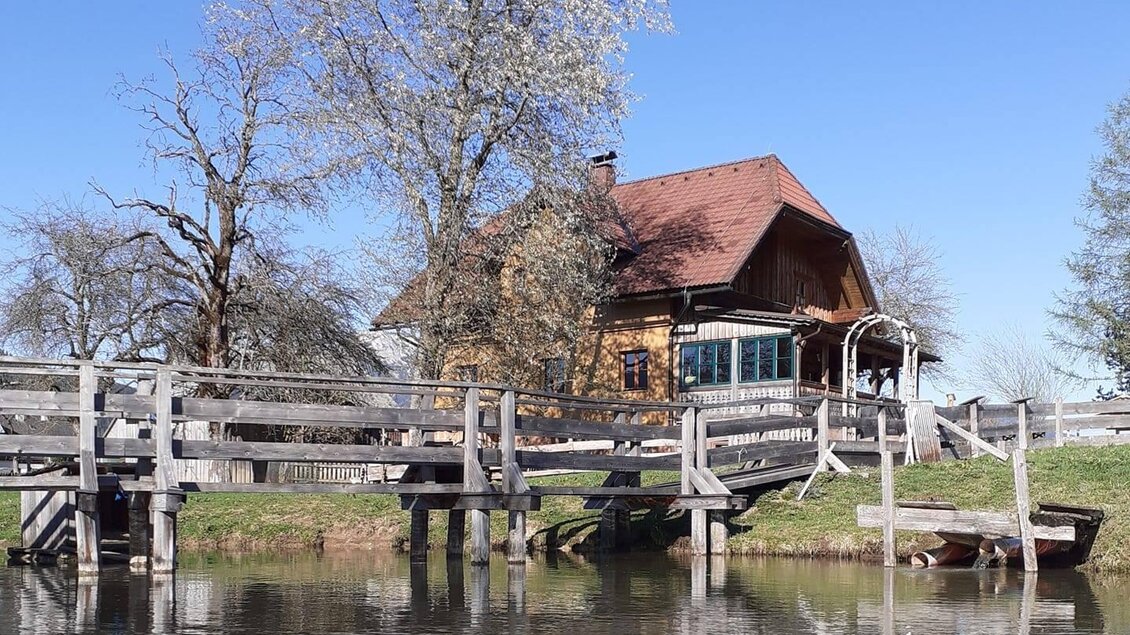 Ein malerisches Haus am Wasser mit einer Holzbrücke. Im Hintergrund sind blühende Bäume und ein klarer blauer Himmel zu sehen.