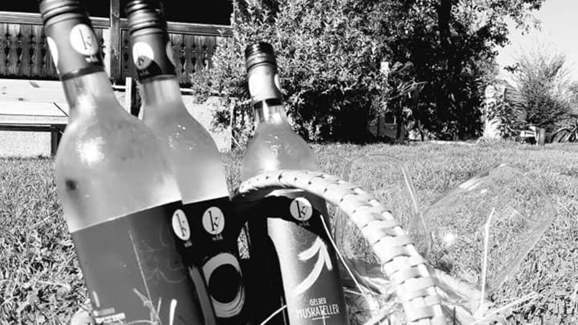 Ein geflochtener Korb mit Flaschen steht auf dem Gras vor einem Holzhaus. Im Hintergrund sind Bäume und ein klarer Himmel sichtbar.