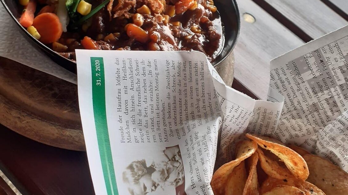 Ein köstliches Gericht in einer Pfanne mit Gemüse und einer Beilage aus Kartoffelchips. Das Essen ist auf einer Holztischplatte angerichtet, umgeben von einer Zeitungsunterlage.
