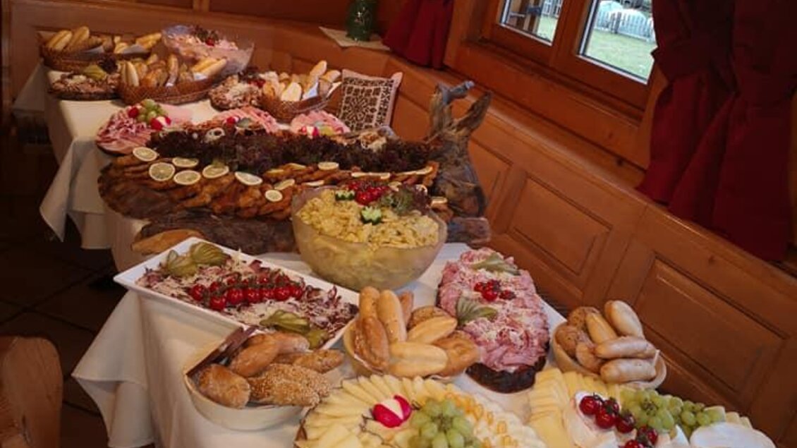 Ein reichhaltiges Büfett mit verschiedenen Leckereien, darunter Kuchen, Obst und Brötchen. Der Tisch ist in einem gemütlichen Raum mit Holzverkleidung gedeckt.