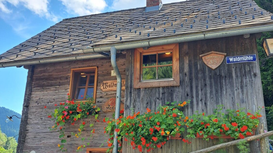 Ein rustikales Holzhaus an einem Hang. Die Umgebung ist von Bäumen und einem Weg gesäumt. | © Tourismusverband Oststeiermark