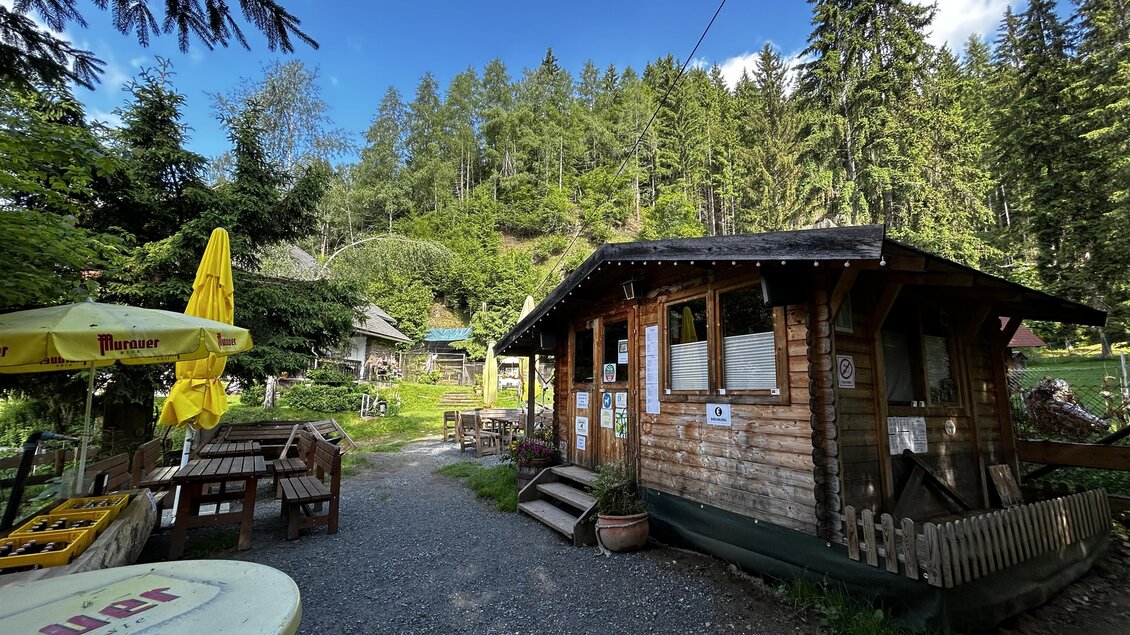 Eine gemütliche Holzblockhütte umgeben von Bäumen und einer ruhigen Landschaft. Im Vordergrund sind Sitzgelegenheiten und Sonnenschirme zu sehen. | © Tourismusverband Murau