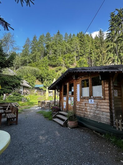 A cozy wooden log cabin surrounded by trees and a tranquil landscape. In the foreground, seating and umbrellas can be seen. | © Tourismusverband Murau
