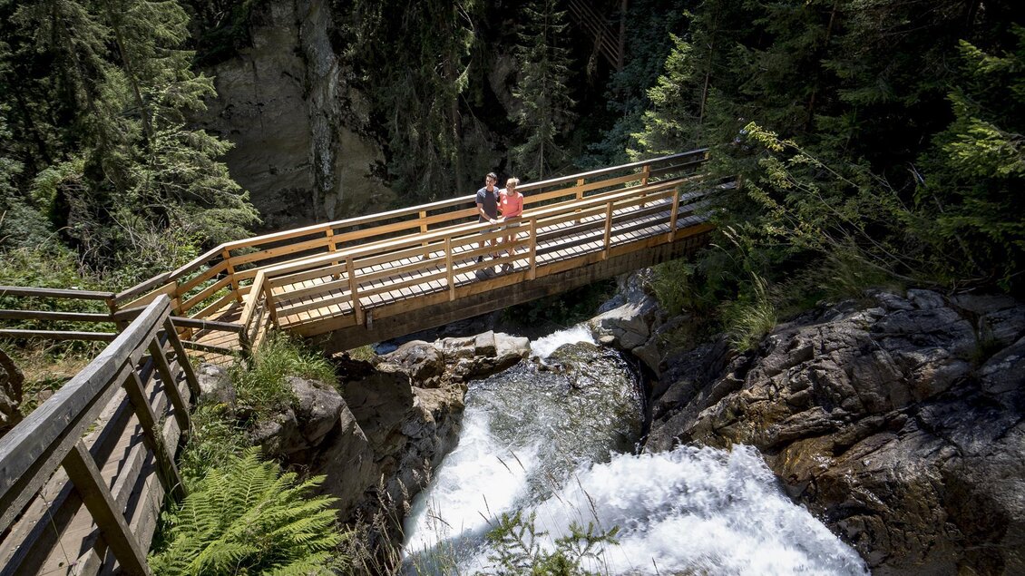 Eine Holzbrücke über einen rauschenden Fluss inmitten eines Waldgebiets. Zwei Personen stehen auf der Brücke und genießen die Aussicht. | © Holzwelt Murau