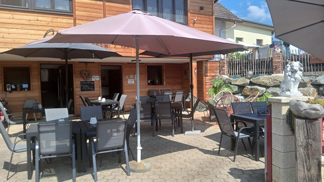 Ein gemütlicher Außenbereich mit Tischen und Stühlen unter Sonnenschirmen. Im Hintergrund ist ein Holzgebäude sichtbar. | © Sarah Windisch