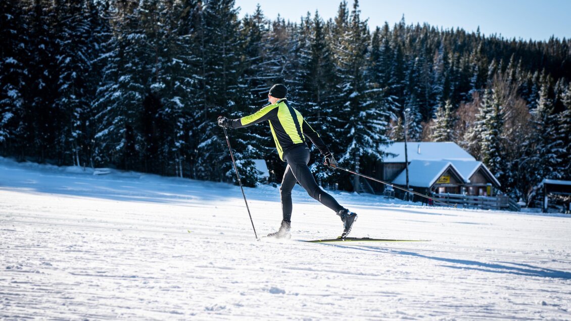 Ein Langläufer in einem neongelben Outfit gleitet durch eine schneebedeckte Landschaft. Im Hintergrund sind Tannenbäume und eine kleine Hütte zu sehen. | © Oststeiermark Tourismus