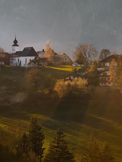 Eine malerische Landschaft mit einem Dorf auf einem Hügel. Die Sonne scheint sanft auf die Häuser und die umliegenden Bäume. | © Christoph Lukas