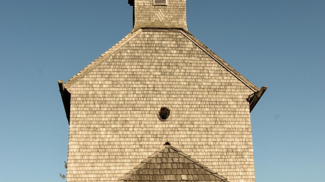 Eine holzverkleidete Kirche mit einem runden Dach und einem Kreuz an der Spitze. Der Himmel ist klar und blau, und die Umgebung ist ländlich.