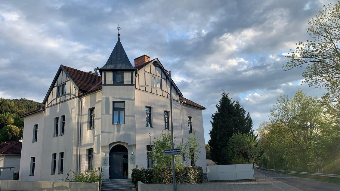 Ein historisches Gebäude mit einem Turm und mehreren Fenstern. Die Umgebung zeigt viel grüne Natur und einen bewölkten Himmel. | © Co:/mikejohann