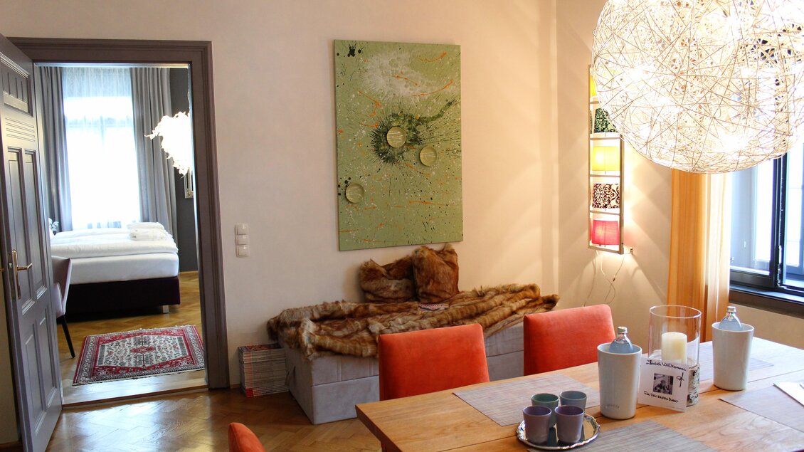 Ein modernes Esszimmer mit einem Holztisch und orangefarbenen Stühlen. Im Hintergrund ist ein Schlafzimmer mit einem großen Bett und einem dekorativen Kunstwerk an der Wand zu sehen. | © Co:/mikejohann