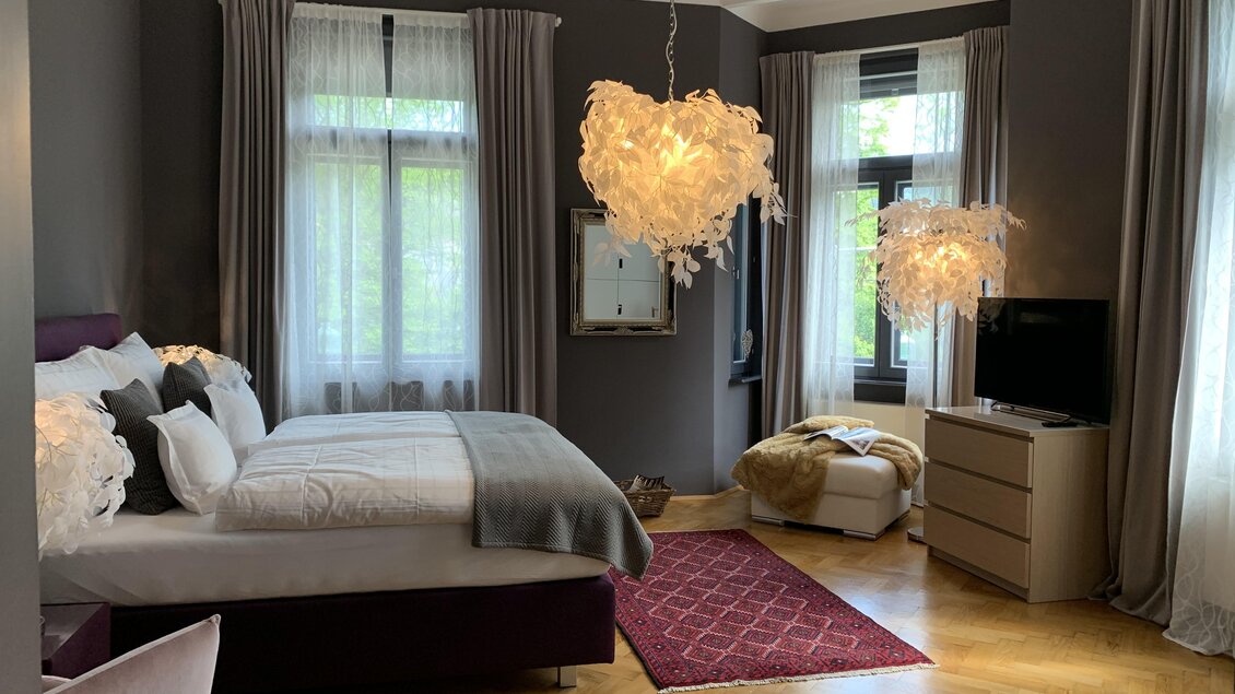 Ein modernes Schlafzimmer mit dunklen Wänden und Holzfußboden. Es gibt einen großen Bett, stilvolle Beleuchtung und einen Fernseher. | © Co:/mikejohann