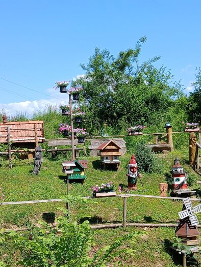 A colorful garden with many birdhouses and green plants. In the background, there are trees and a clear sky. | © Maria Fink