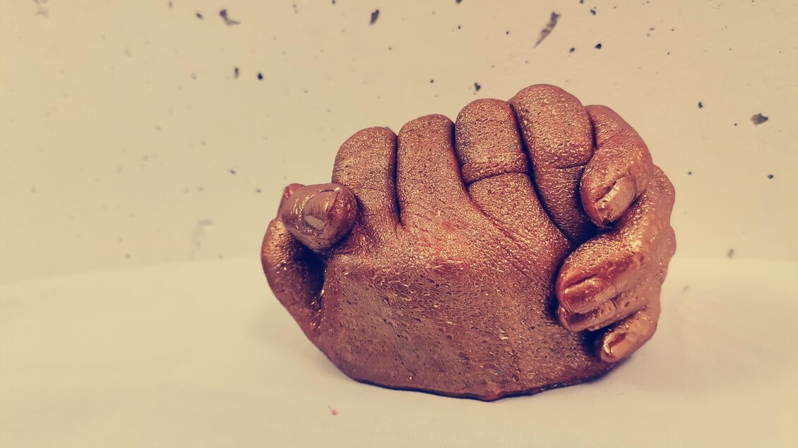 Eine stilisierte Skulptur von zwei miteinander verflochtenen Händen in bronzefarbenem Finish. Die Hände scheinen sich fest zu umarmen und symbolisieren Zusammenarbeit oder Einheit. | © Julia Hauck 