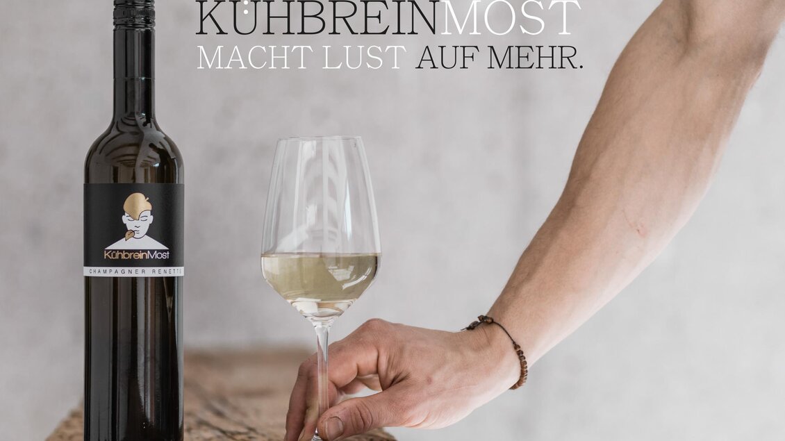 Eine Flasche Kuhbreinmost steht neben einem halb vollen Weinglas auf einem Tisch. Ein Arm mit einem Armband hält das Glas und suggeriert Genuss und Eleganz. | ©  Kühbrein