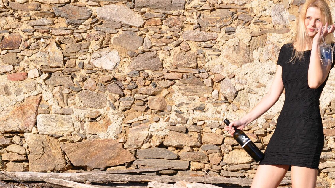 Eine Frau in einem schwarzen Kleid steht vor einer Steinhintergrund. Sie hält eine Flasche Sekt und ein Glas in der Hand. | ©  Kühbrein