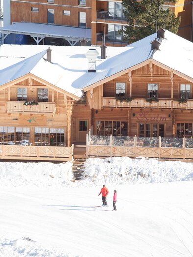 A charming wooden house in a snow-covered landscape. Two people are walking on the snow in front of the building. | © Tourismusverband Murau