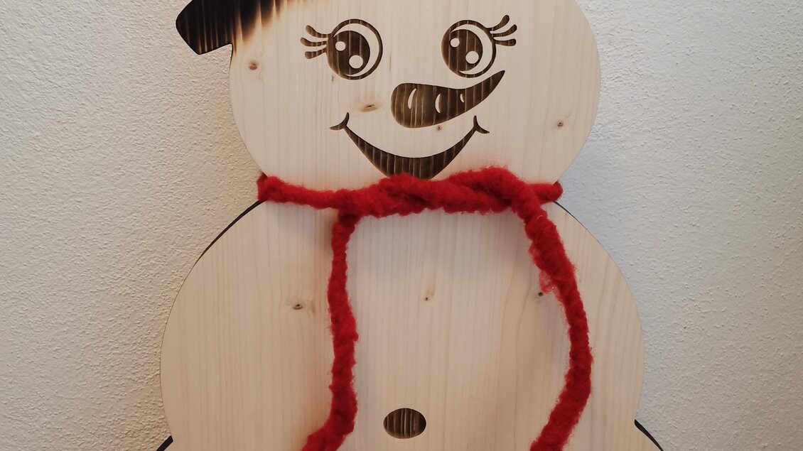 Eine Holzfigur eines Schneemanns mit einem Zylinderhut und einem roten Schal. Der Schneemann hat ein freundliches Gesicht mit lächelnden Augen. | © KM Lasergravuren