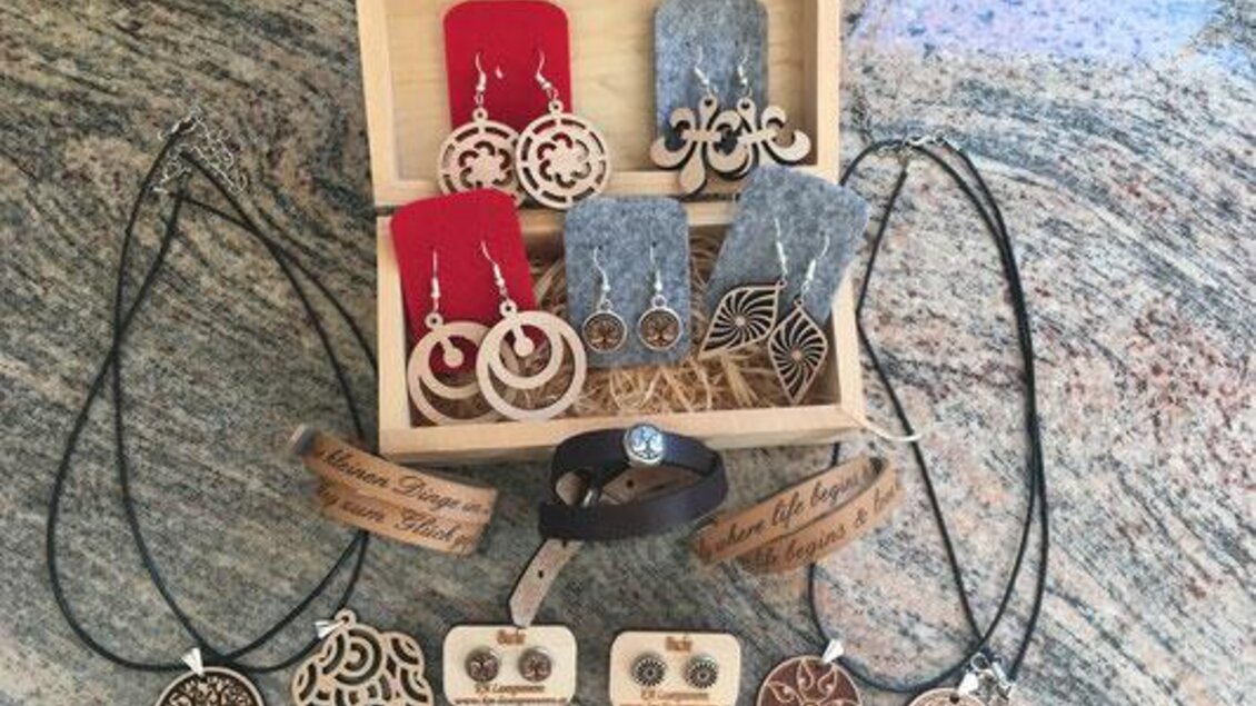 Eine Auswahl von handgemachtem Schmuck, einschließlich Ohrringen und Halsketten, präsentiert in einer Holzkiste. Der Schmuck besteht aus Holz und anderen Materialien in verschiedenen Designs und Farben. | © KM Lasergravuren