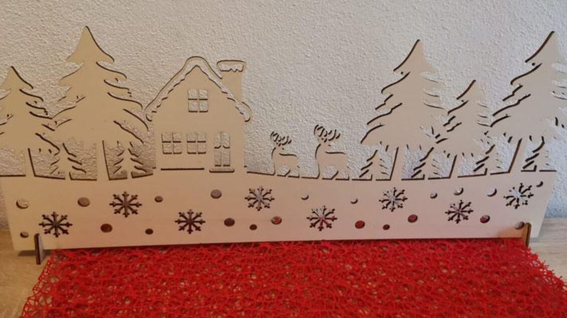 Eine dekorative Holzschnitzerei mit Tannenbäumen, einem Haus und Rentieren. Im Vordergrund sind Schneeflocken zu sehen, die eine winterliche Stimmung erzeugen. | © KM Lasergravuren